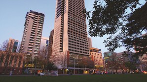 Un immeuble moderne de grande hauteur, doté d’une architecture originale, entouré d’un parc arboré et offrant une vue sur un ciel dégagé.