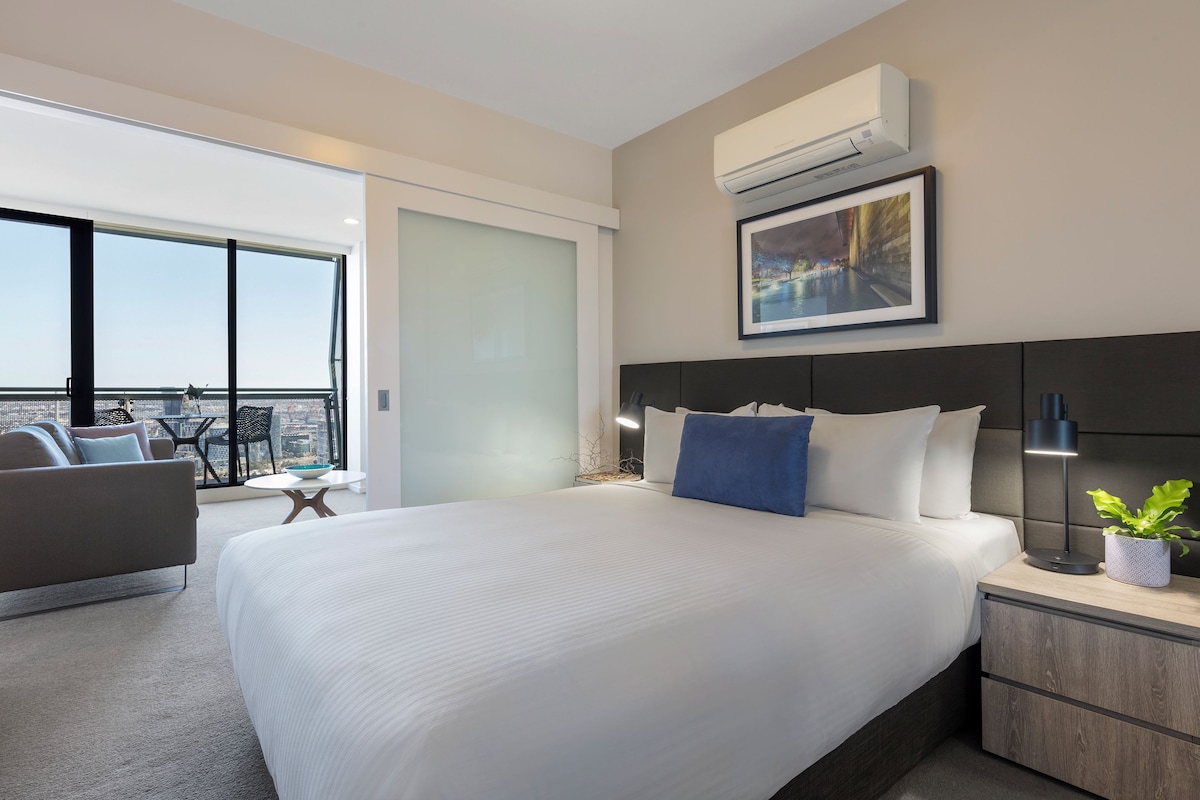Une chambre d’hôtel moderne avec un grand lit, une table de chevet avec une lampe, une petite table d’appoint, un canapé et une grande fenêtre donnant sur la ville.