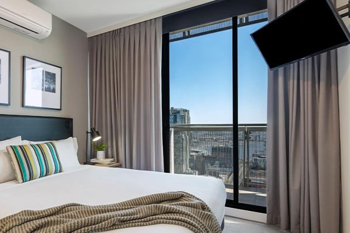 Une chambre d’hôtel avec un grand lit, une table de chevet, une lampe, un téléviseur à écran plat et une fenêtre avec des rideaux.
