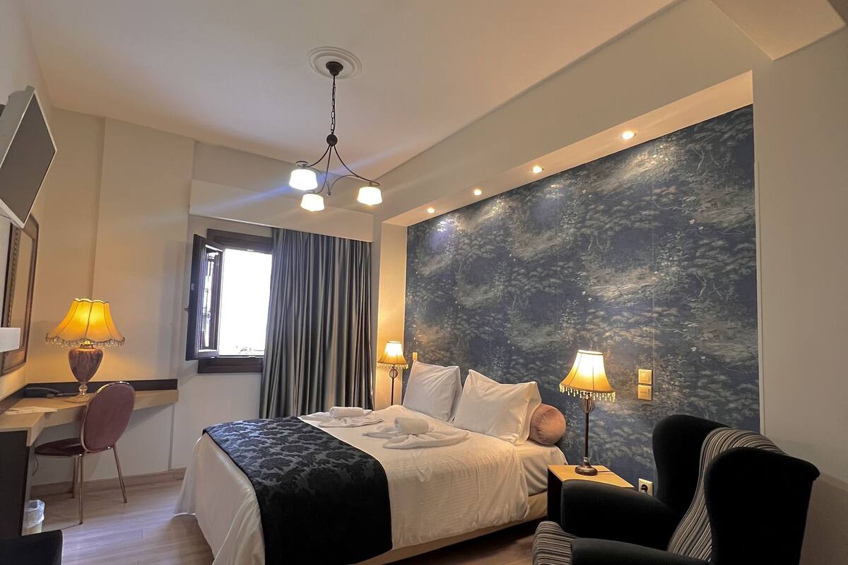 Una habitación de hotel moderna con una cama grande, dos lámparas de noche, un escritorio con una silla, un ventanal con cortinas y un mural en la pared.