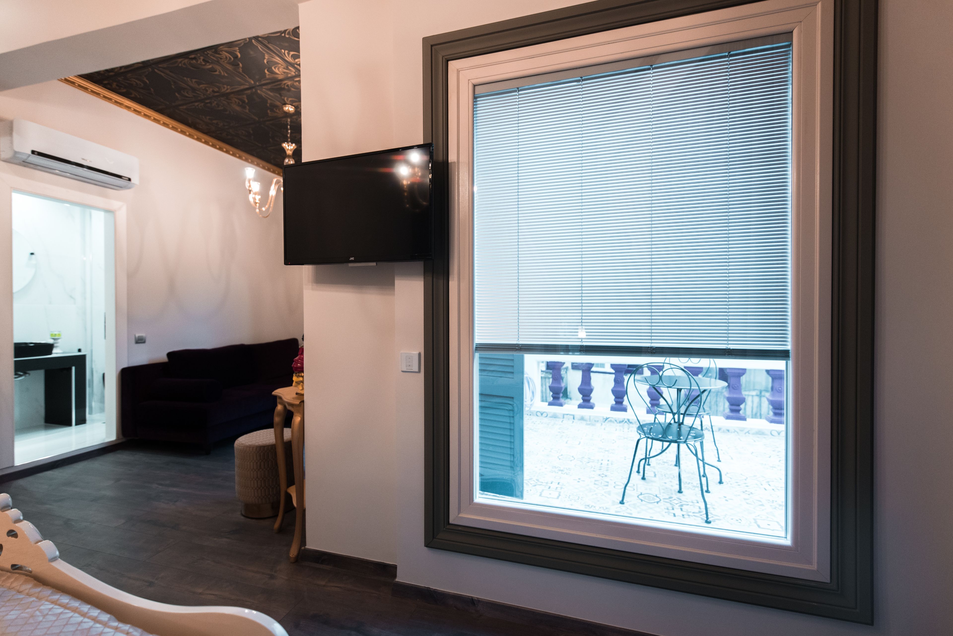 A modern living room with a large window, a flat-screen TV mounted on the wall, and a dining area visible through the window.