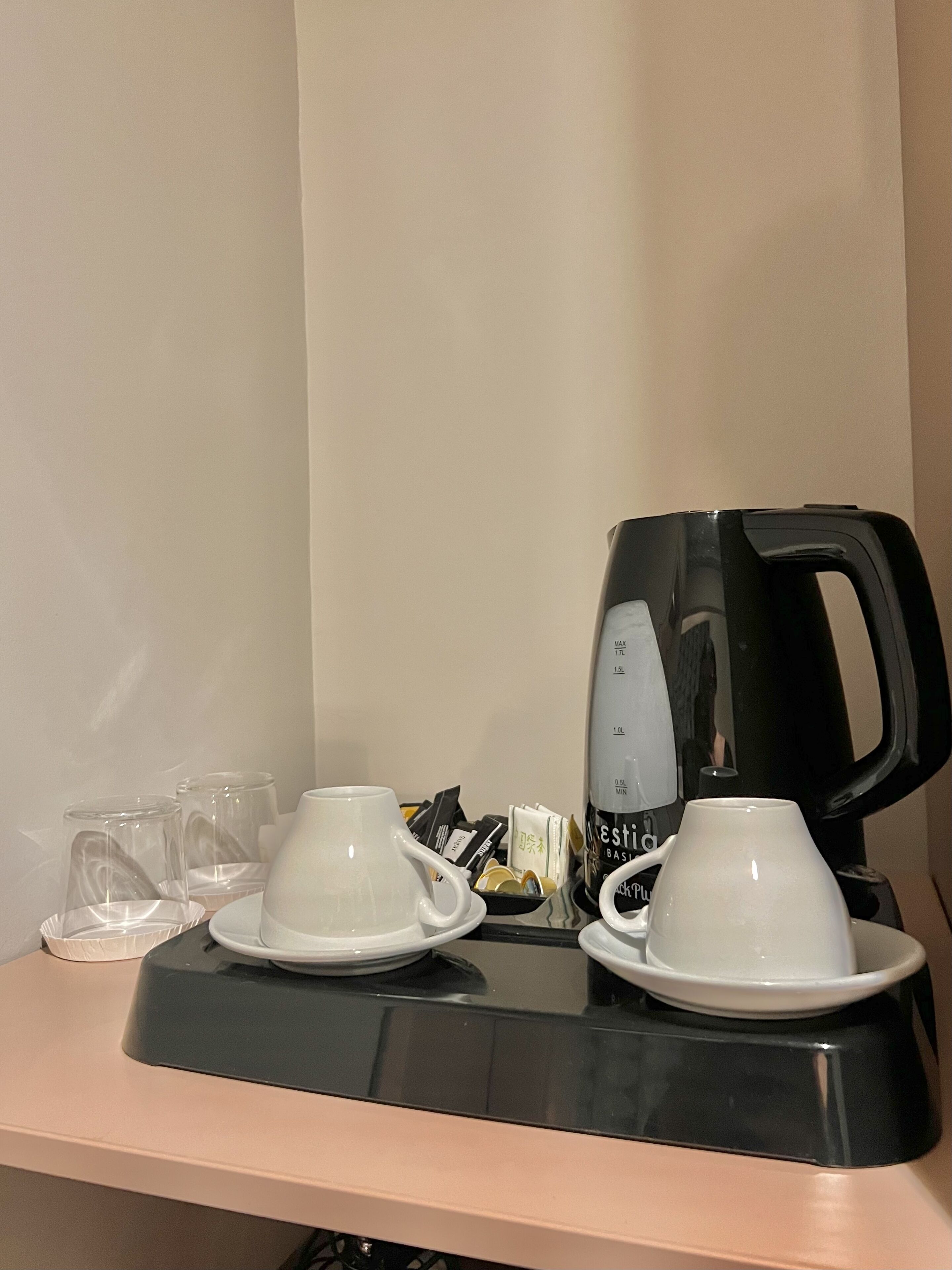 Una estación de café con una cafetera negra, dos tazas blancas y varias tazas de vidrio vacías sobre una encimera color beige.