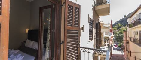 Una calle urbana estrecha flanqueada por edificios, un balcón con barandilla metálica y una cama con ropa blanca.