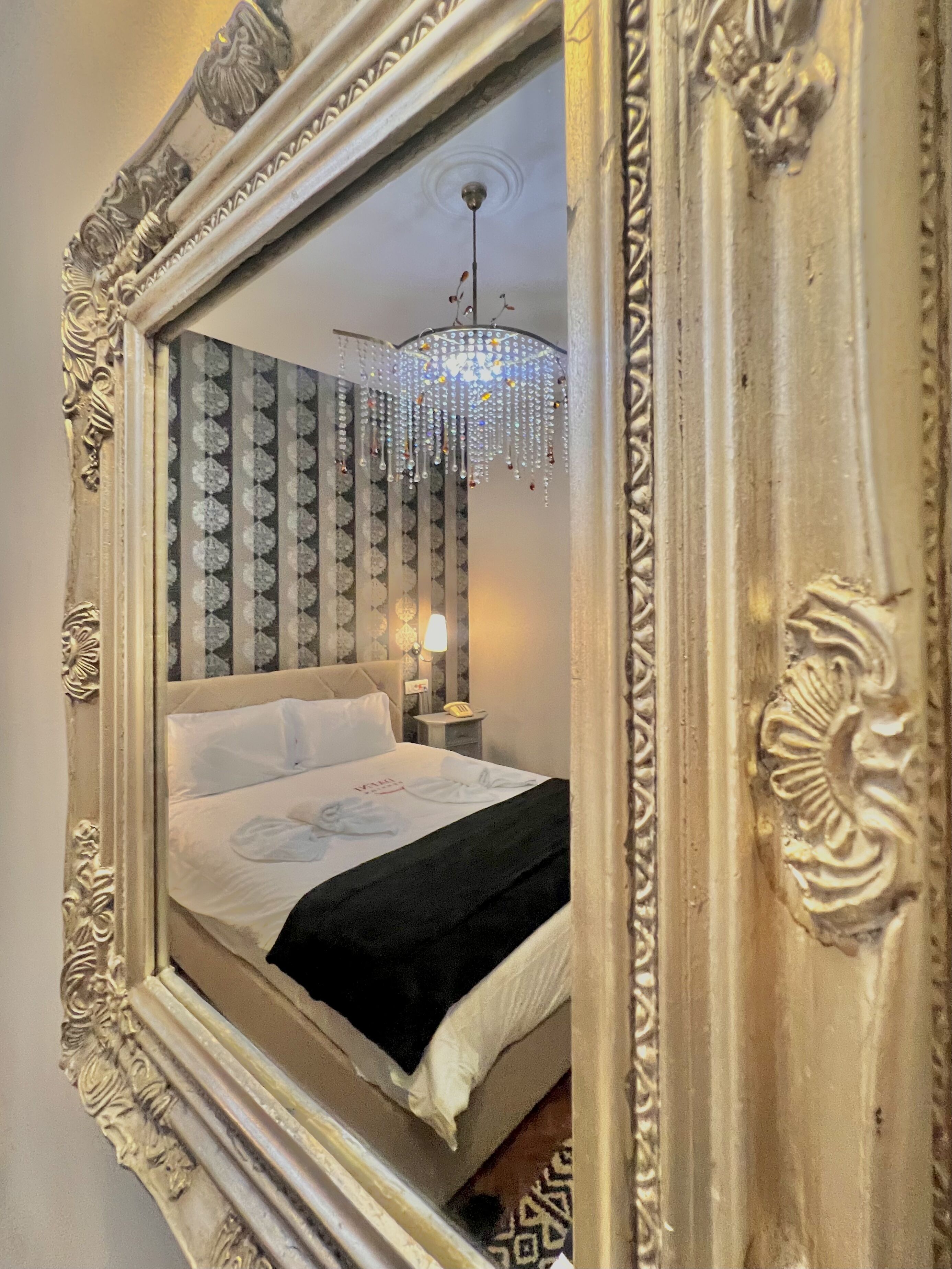 Habitación de hotel con un espejo grande y ornamentado, una cama con ropa de cama blanca, una manta de color negro, un candelabro y un papel tapiz estampado.