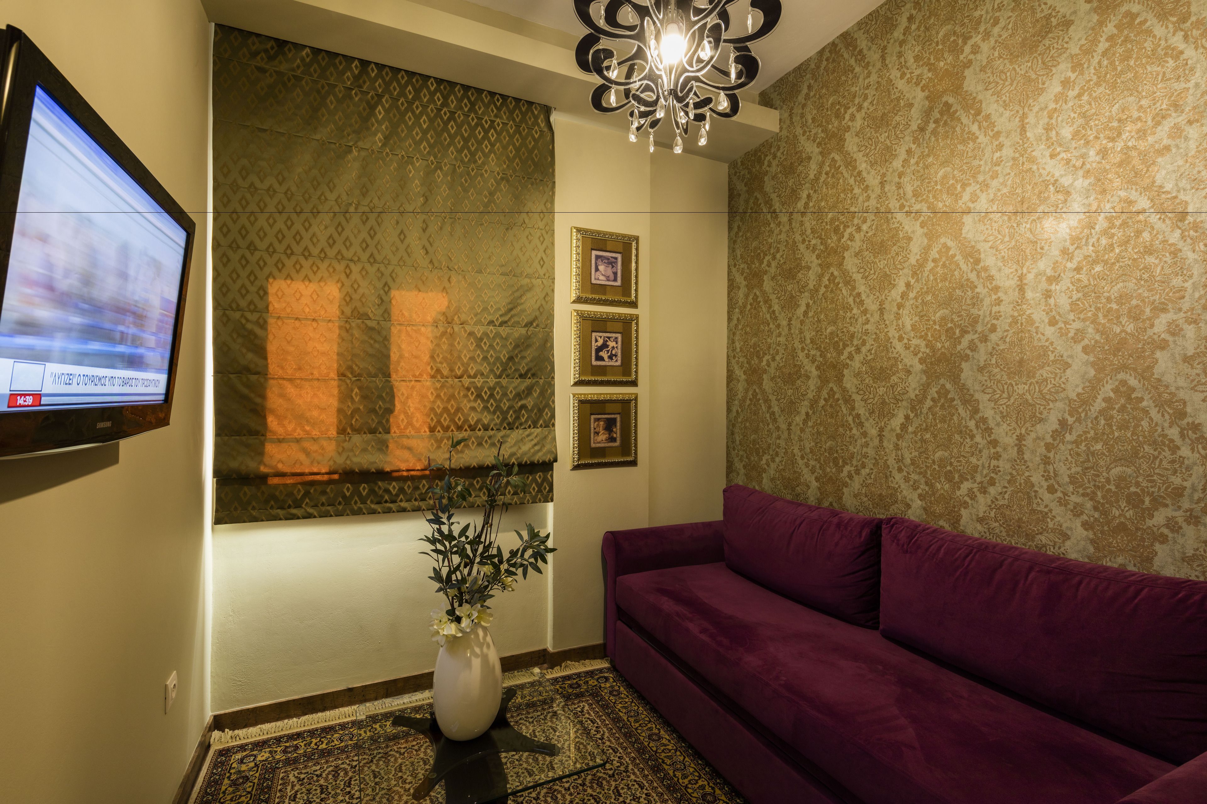 A room with a purple sofa, a wall-mounted TV, a decorative plant, patterned wallpaper, and a chandelier.