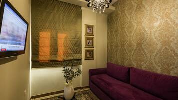 A room with a purple sofa, a wall-mounted TV, a decorative plant, patterned wallpaper, and a chandelier.
