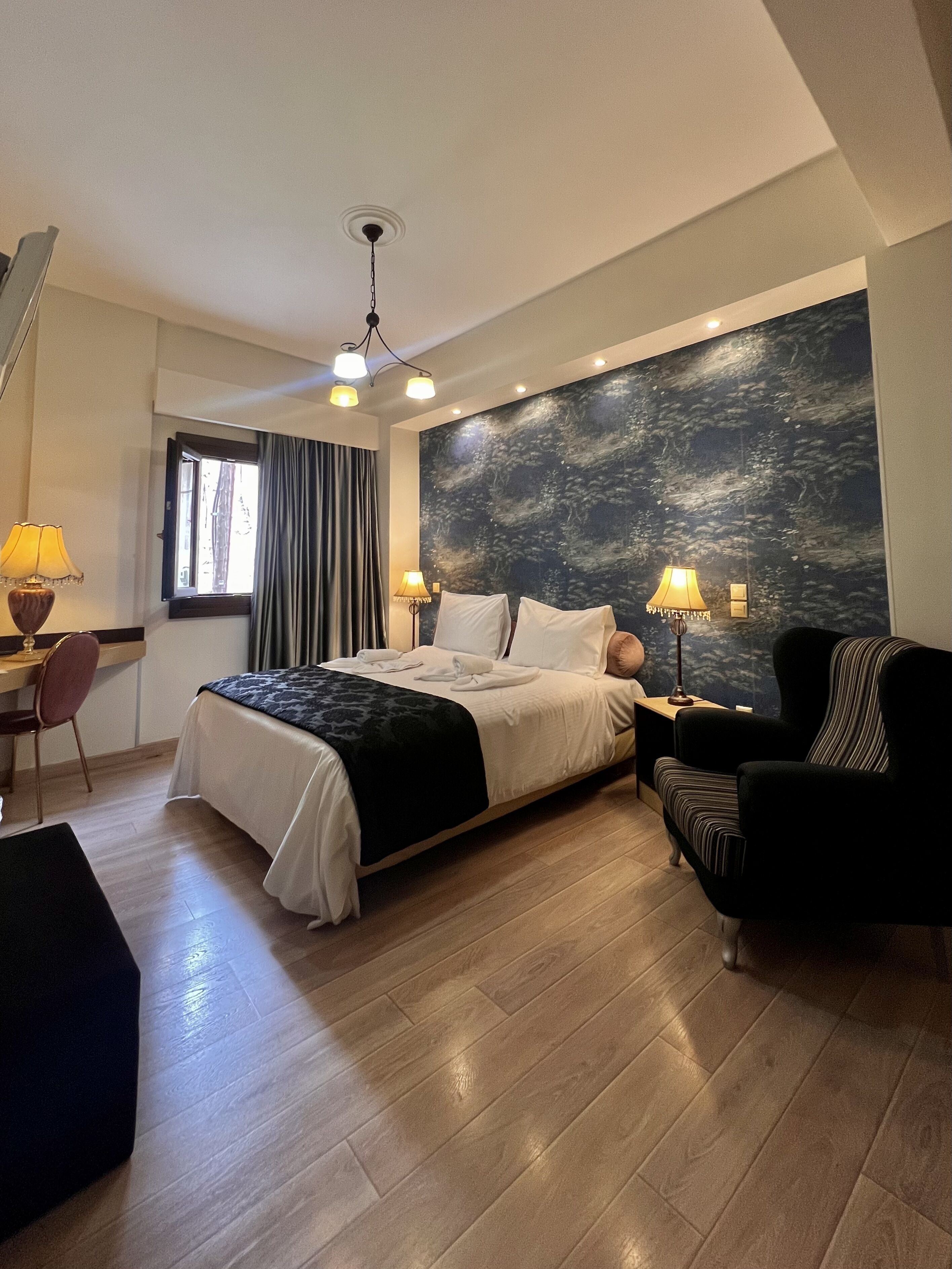 Une chambre d’hôtel comprenant un lit, deux lampes de chevet, un bureau avec une chaise, une grande fenêtre avec des rideaux et un mur orné d’une fresque à motifs.