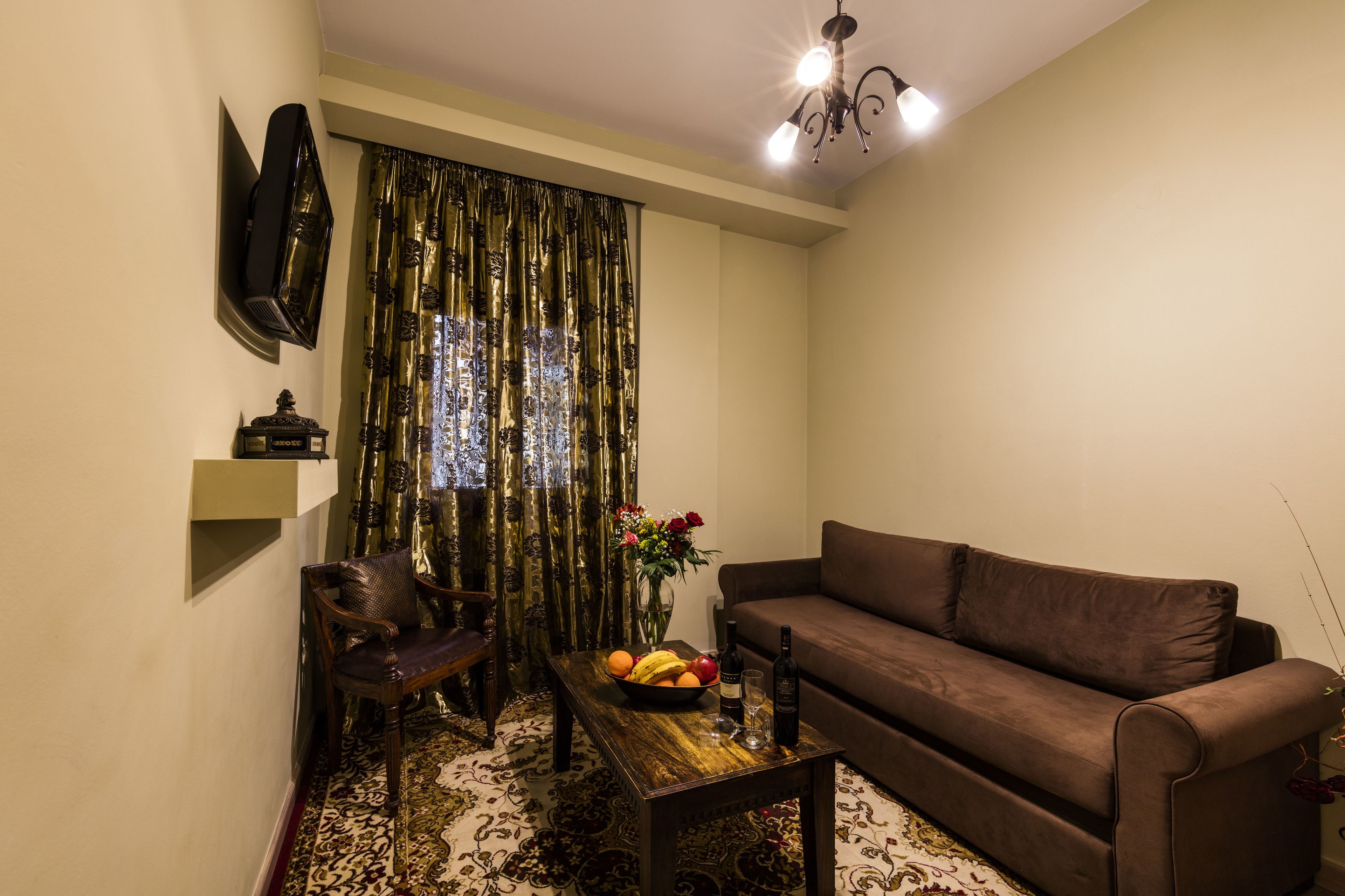 Una sala de estar con un sofá marrón, una mesa de centro de madera, una silla, un televisor montado en la pared y una cortina decorativa.