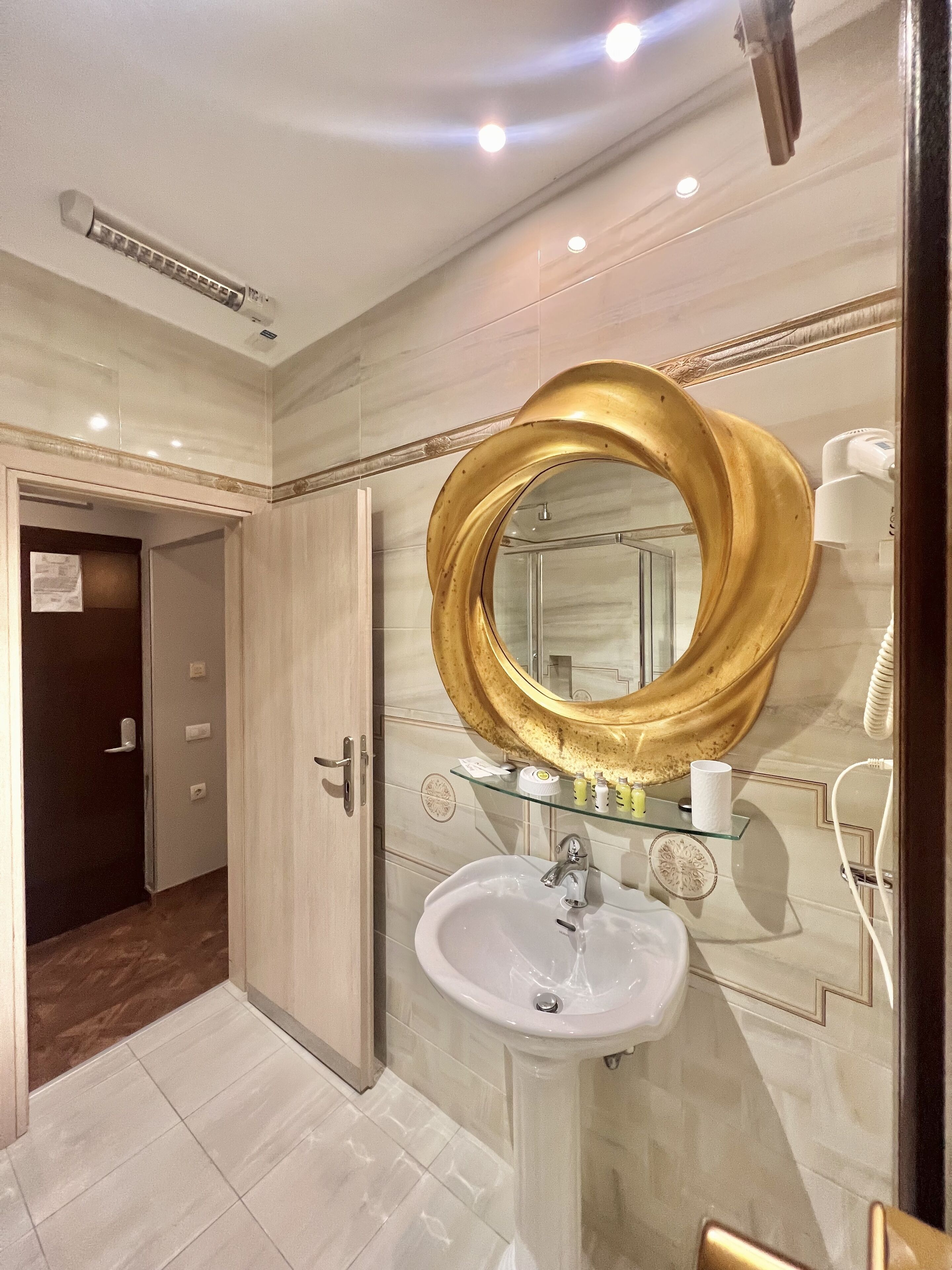A bathroom with a large golden mirror, a white sink, and a glass shelf with toiletries.