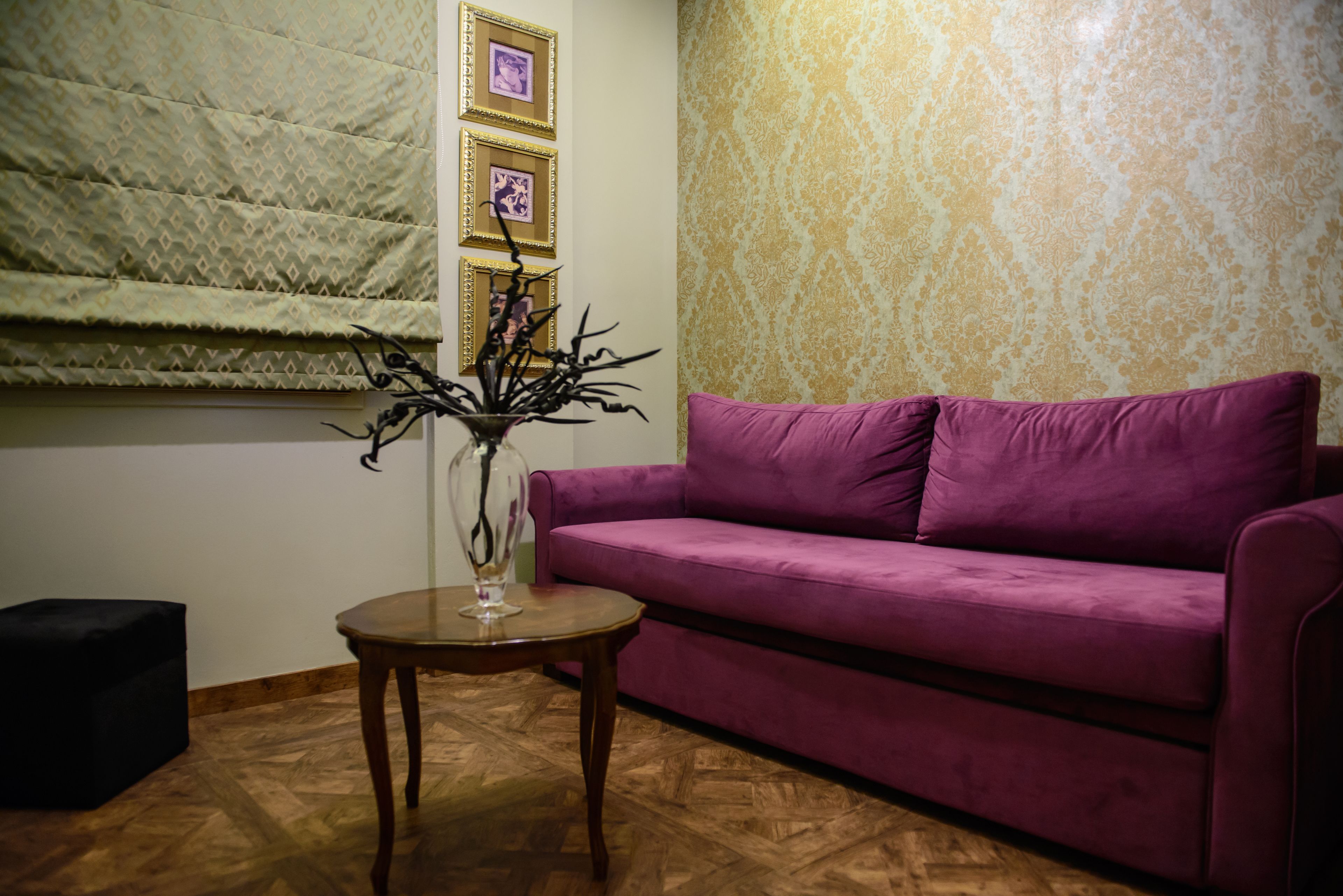 A room with a purple sofa, a small wooden table with a vase, patterned curtains, and a patterned wall.