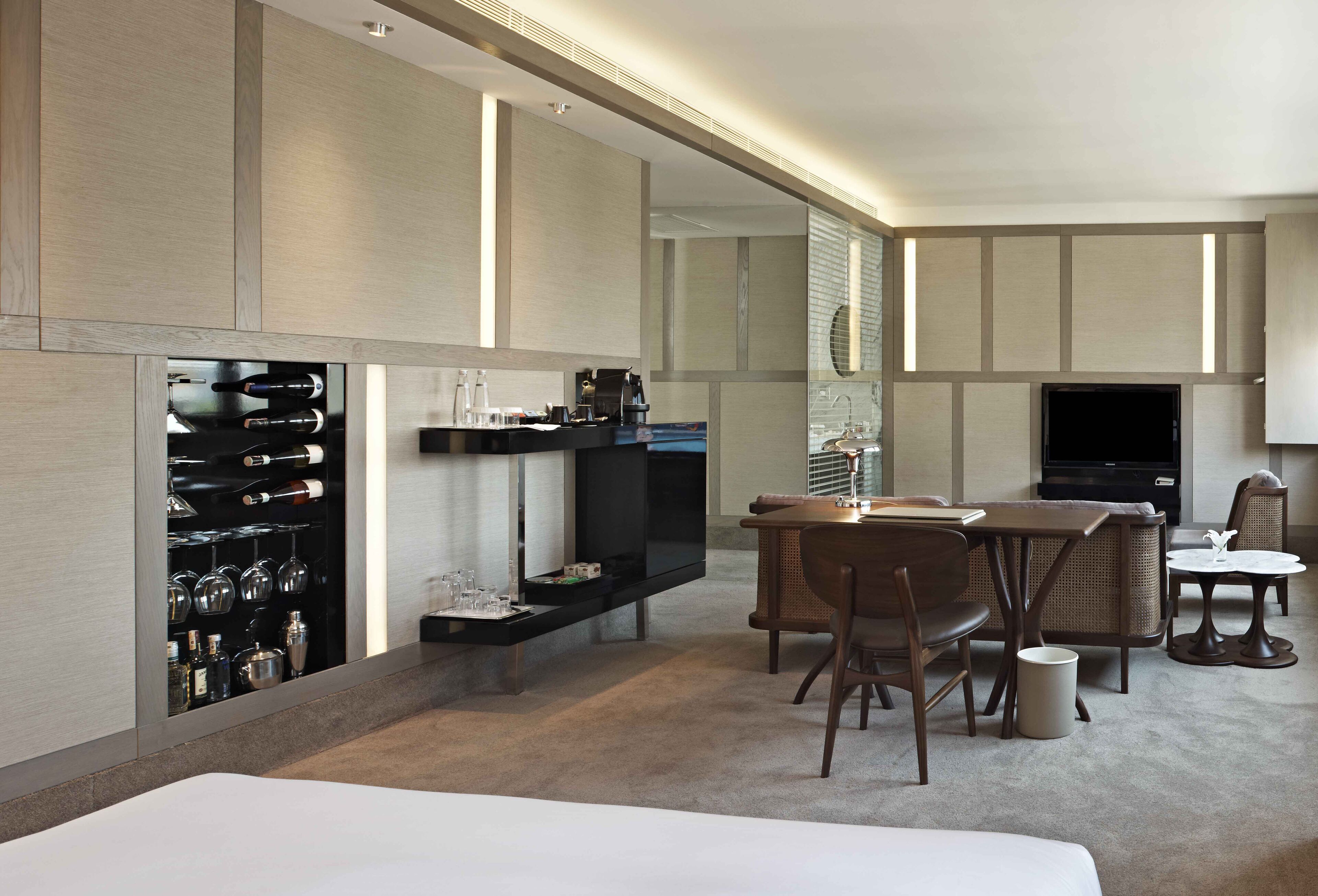 A modern hotel room with a flat-screen TV, a minibar, a dining area with wicker furniture, and a large window.