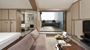 Ein modernes Hotelzimmer mit Balkon, einer Sitzecke, einem Esstisch mit Früchten und Gläsern sowie einem Fernseher.