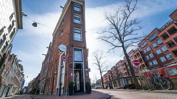 A tall red brick building with a modern glass entrance, a no parking sign, and bicycles parked outside. - Linden Hotel (Amsterdam)