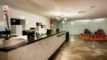 A modern reception area with a marble counter, two black chairs, a potted plant, and a seating area with red and grey chairs.