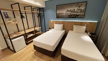 A hotel room with two beds, a desk, a small refrigerator, and a wall-mounted shelf with a telephone and a clock.