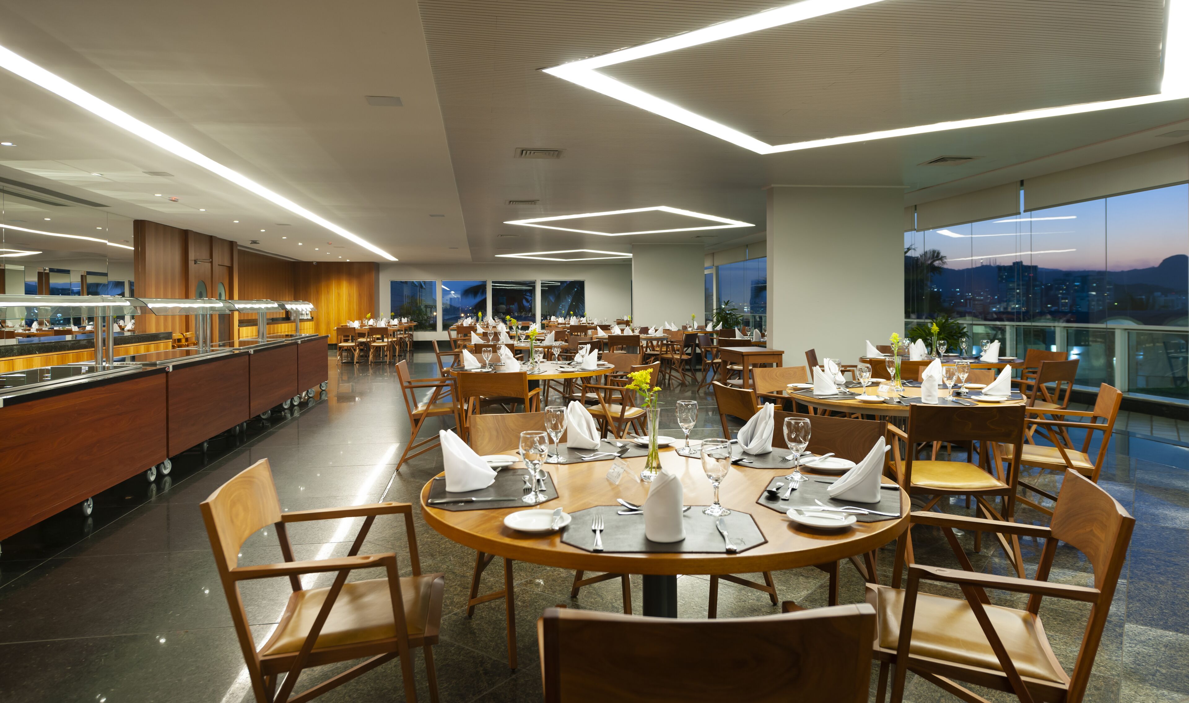 A spacious dining area with multiple tables and chairs, a buffet counter, and large windows offering a city view.