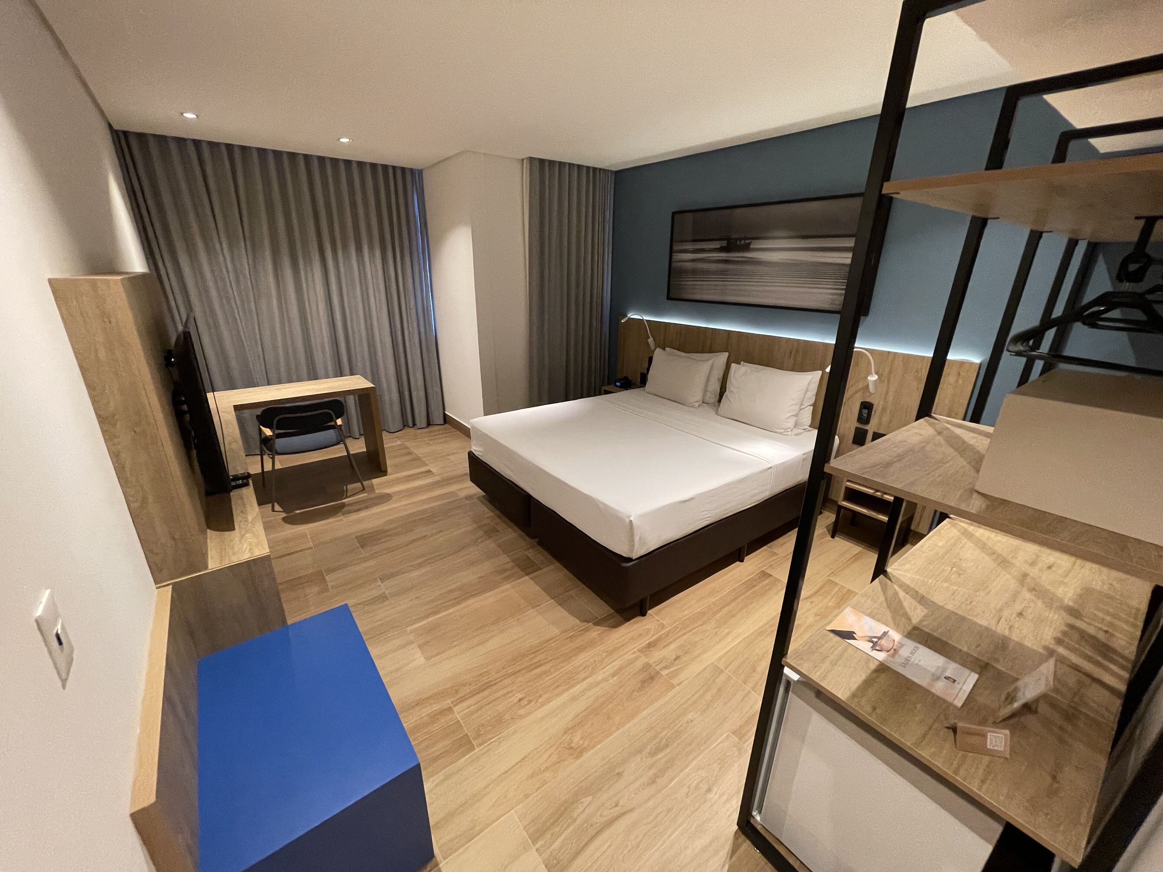 A modern hotel room with a large bed, a desk, a chair, and a wardrobe.