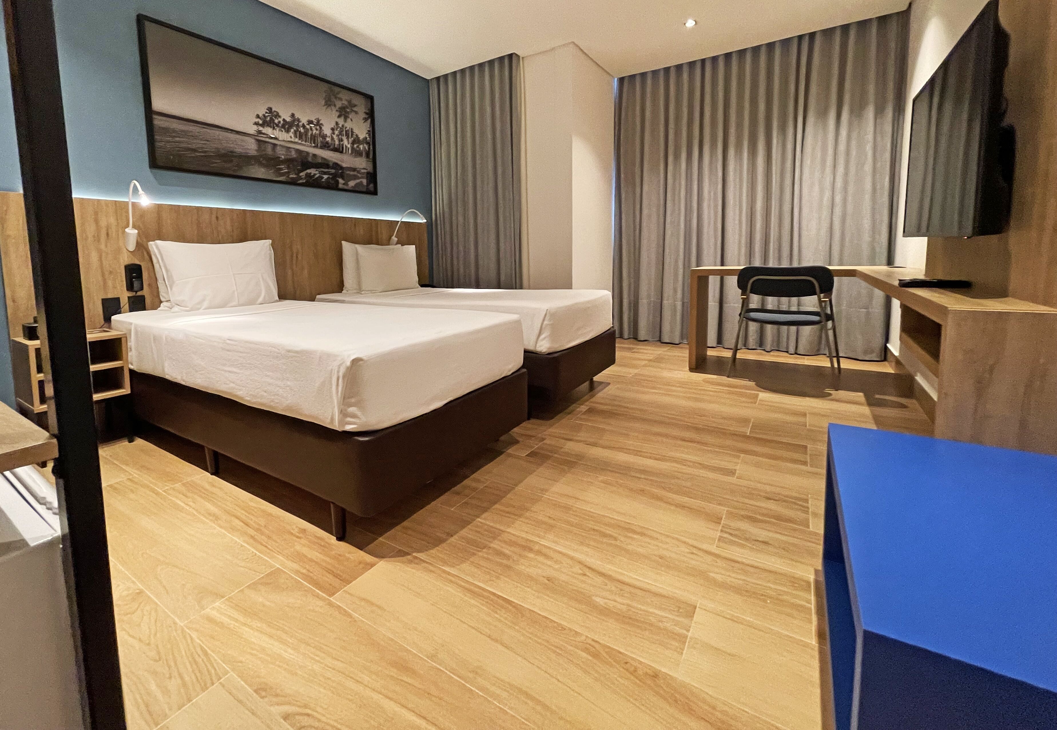 Quarto de hotel com duas camas, uma escrivaninha, uma cadeira, uma televisão e um quadro na parede.