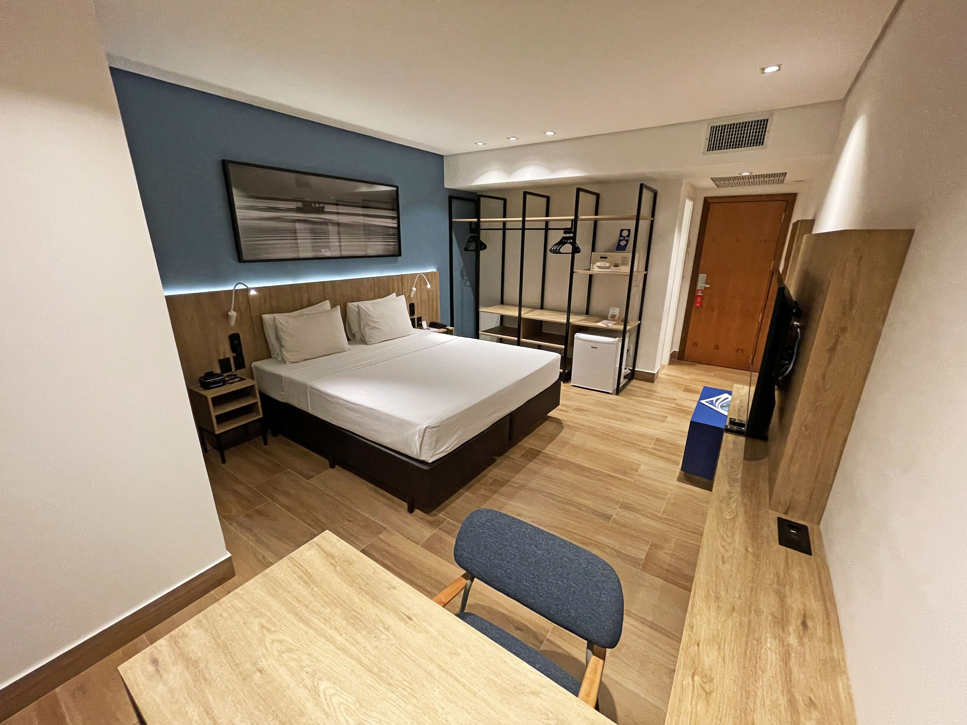Una habitación de hotel moderna con una cama grande, un escritorio y una silla. Hay un refrigerador pequeño y un televisor sobre un mueble.