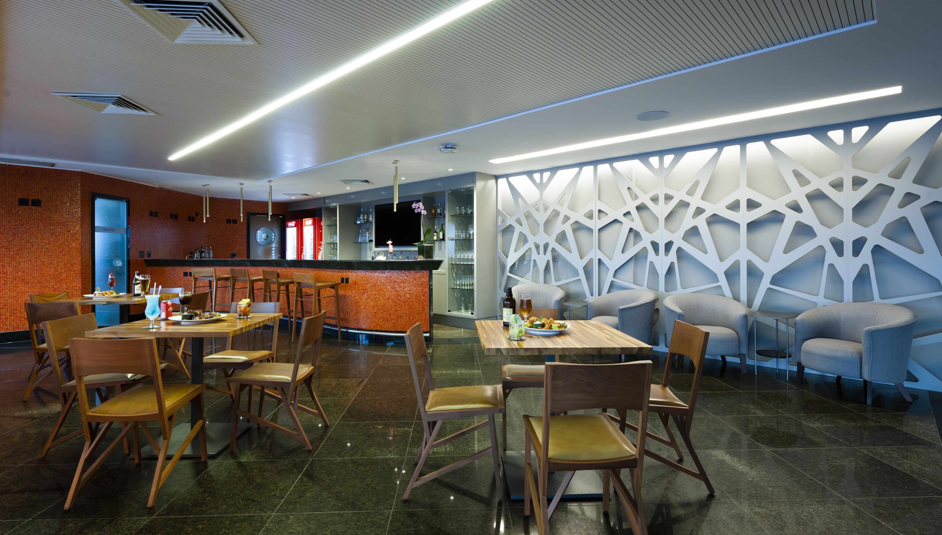 A modern restaurant with wooden tables and chairs, a bar area, and a decorative wall with geometric patterns.