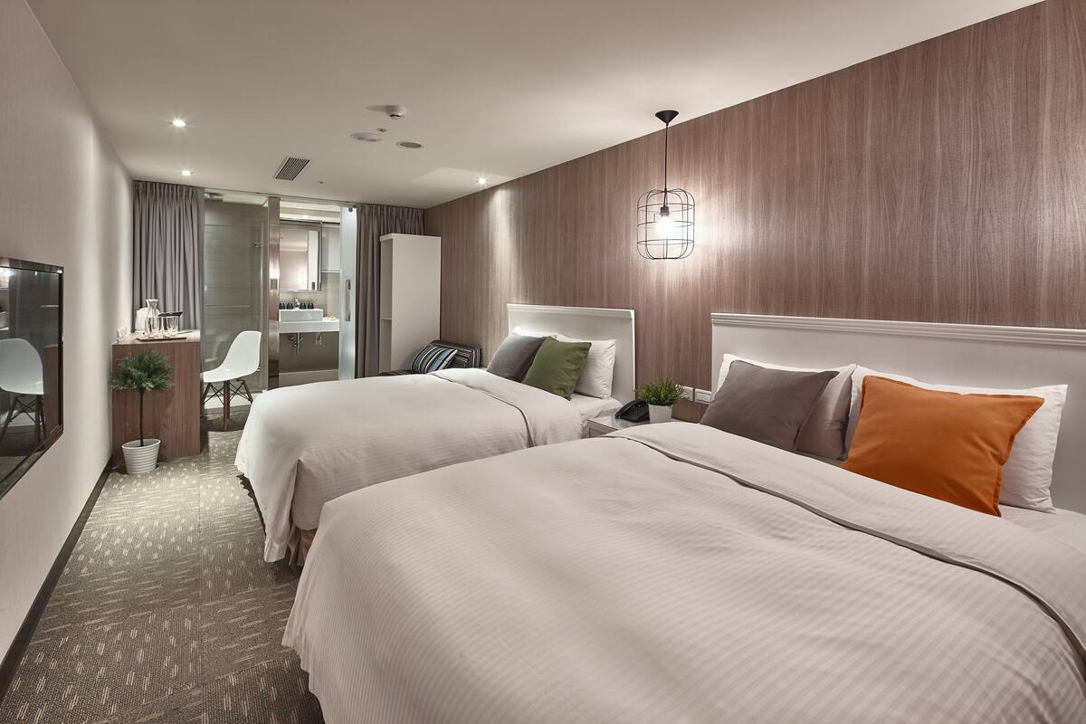 A hotel room with two beds, a desk, a chair, a small plant, and a bathroom visible through a glass partition.