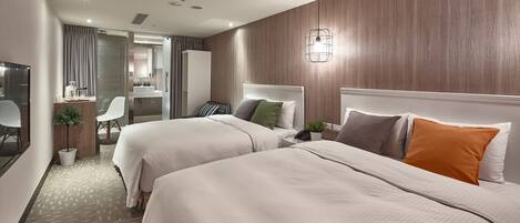 Une chambre d’hôtel avec deux lits, un bureau, une chaise, une petite plante et une salle de bain visible à travers une paroi en verre.