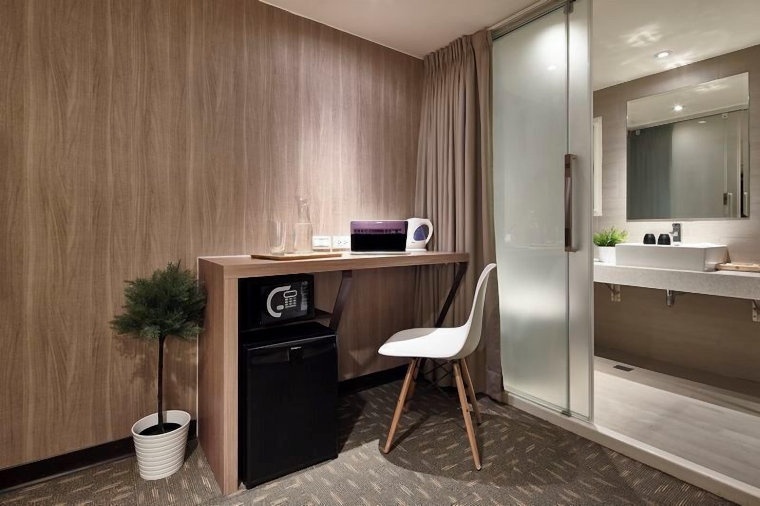 A compact hotel room with a wooden desk, a small refrigerator, a white chair, a microwave, and a bathroom with a mirror and sink.
