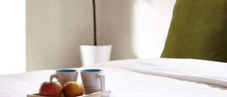 A neatly made bed with a magazine, two apples, and two mugs on a tray. A small potted plant is beside the bed, and a wall decoration reads "c'est la vie".