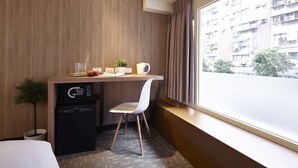 A compact hotel room with a wooden wall, a small desk with a chair, a mini fridge, and a window with a view of a building. - Via Hotel Loft (Taipei)