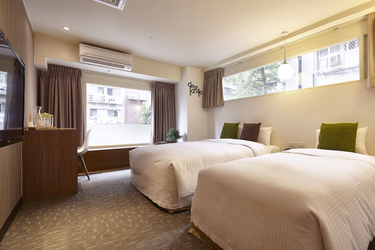 A hotel room with two beds, a desk, and a large window with curtains.