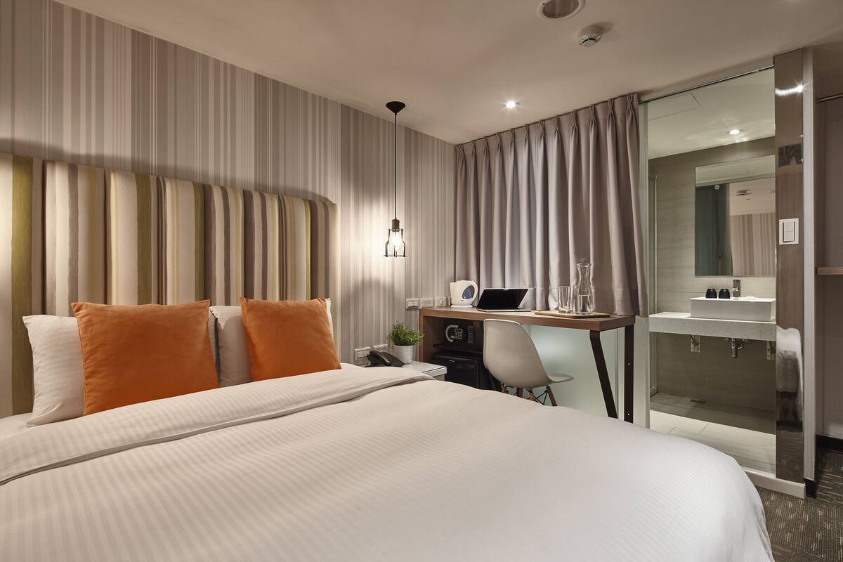 A hotel room with a large bed, a desk with a laptop, a chair, and a bathroom visible through a glass door.