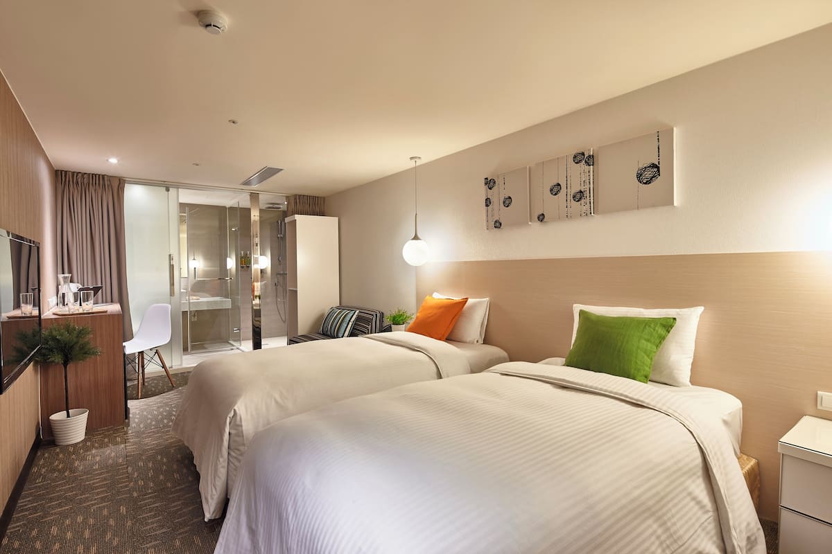 A hotel room with two beds, a desk, a chair, a small plant, and a mirror.