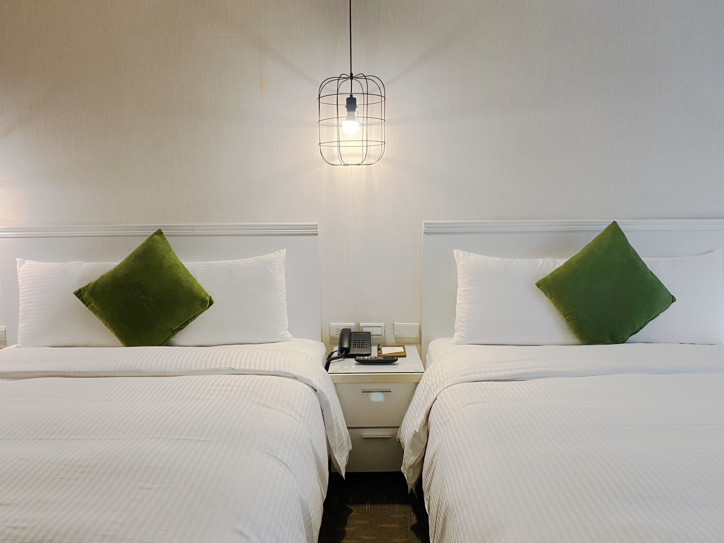 A hotel room with two beds, each with a green pillow, a bedside table with a phone and a lamp, and a hanging light fixture.