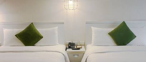 A hotel room with two beds, each with a green pillow, a bedside table with a phone and a lamp, and a hanging light fixture.