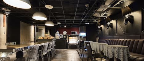 A restaurant interior with a herringbone wooden floor, metal-framed tables, and chairs. There are hanging pendant lights and a counter with a display of food items.