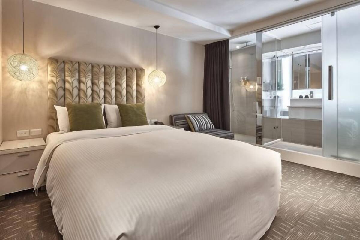 A modern hotel room with a large bed, bedside tables, a glass-enclosed bathroom, and a view of the city.