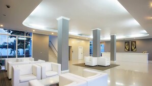 A modern lobby with white sofas, a reception desk, and large windows offering a view of palm trees. - La Vie En Rose Hotel (Chiang Rai)