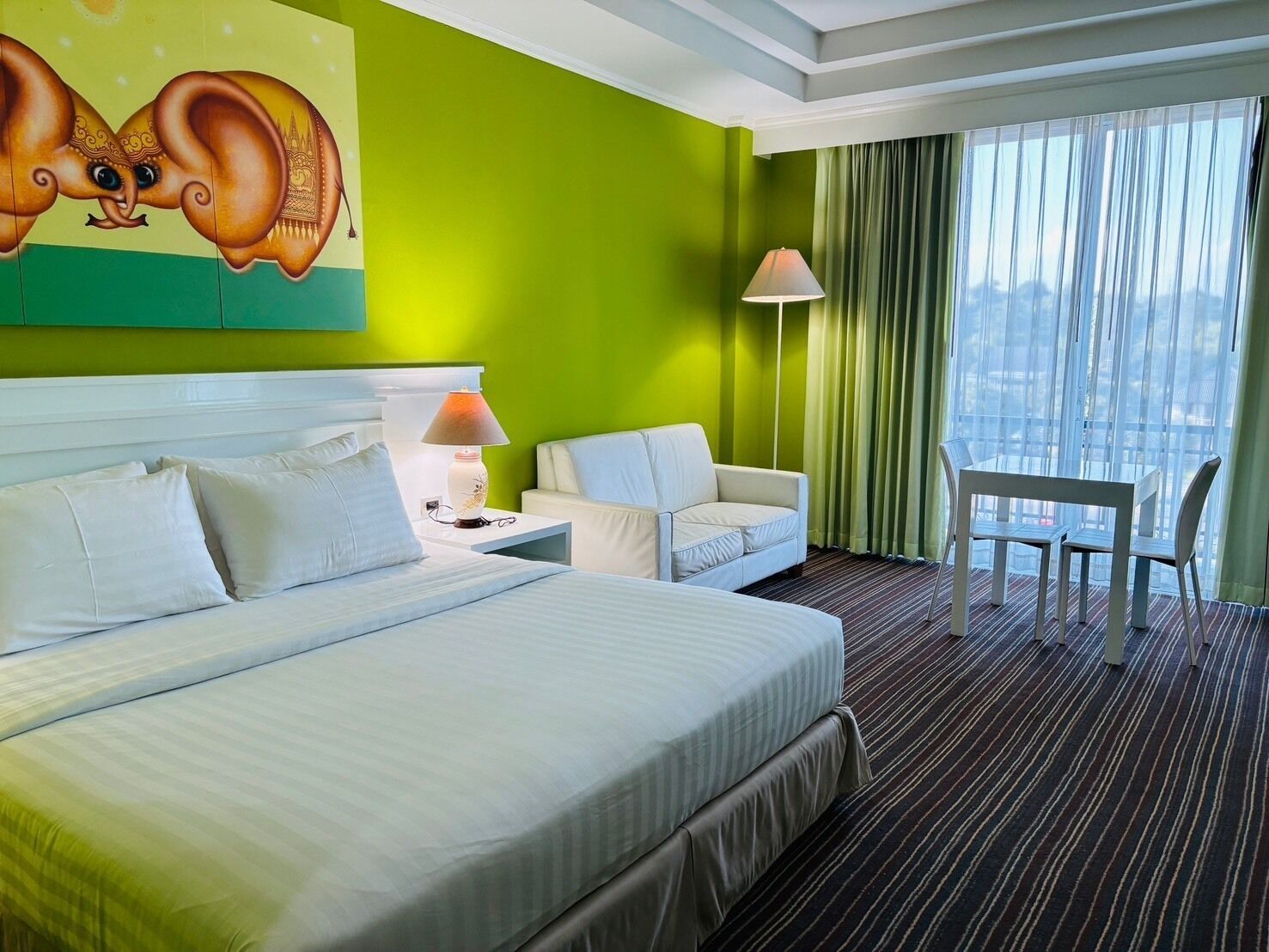 A hotel room with a large bed, a sofa, a small table, and a chair. There is a painting on the wall and a window with curtains.