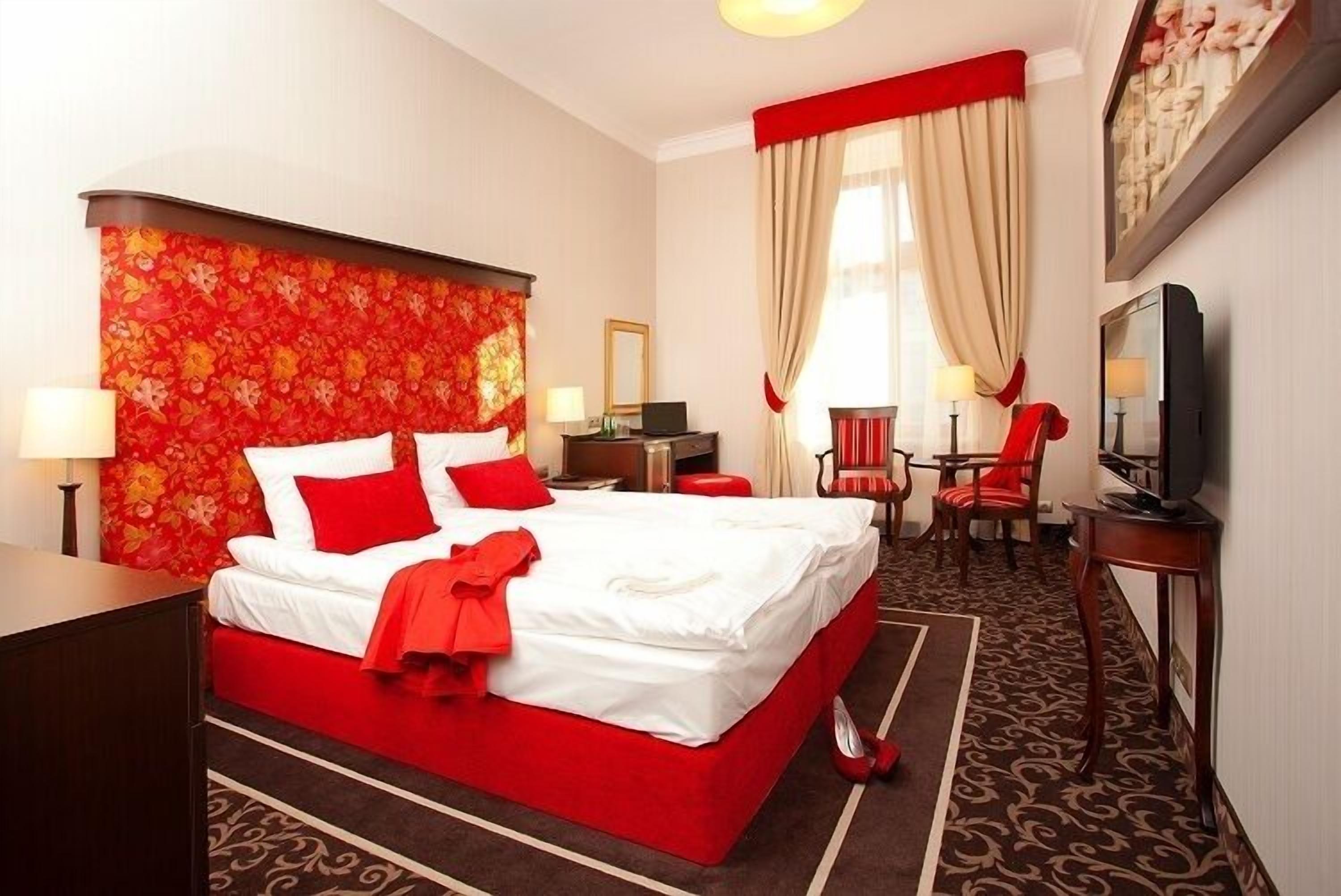 A hotel room with a large bed, red and white bedding, a red throw, a television, a desk with a lamp, and a red chair.