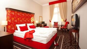 A hotel room with a large bed, red and white bedding, a red throw, a television, a desk with a lamp, and a red chair.