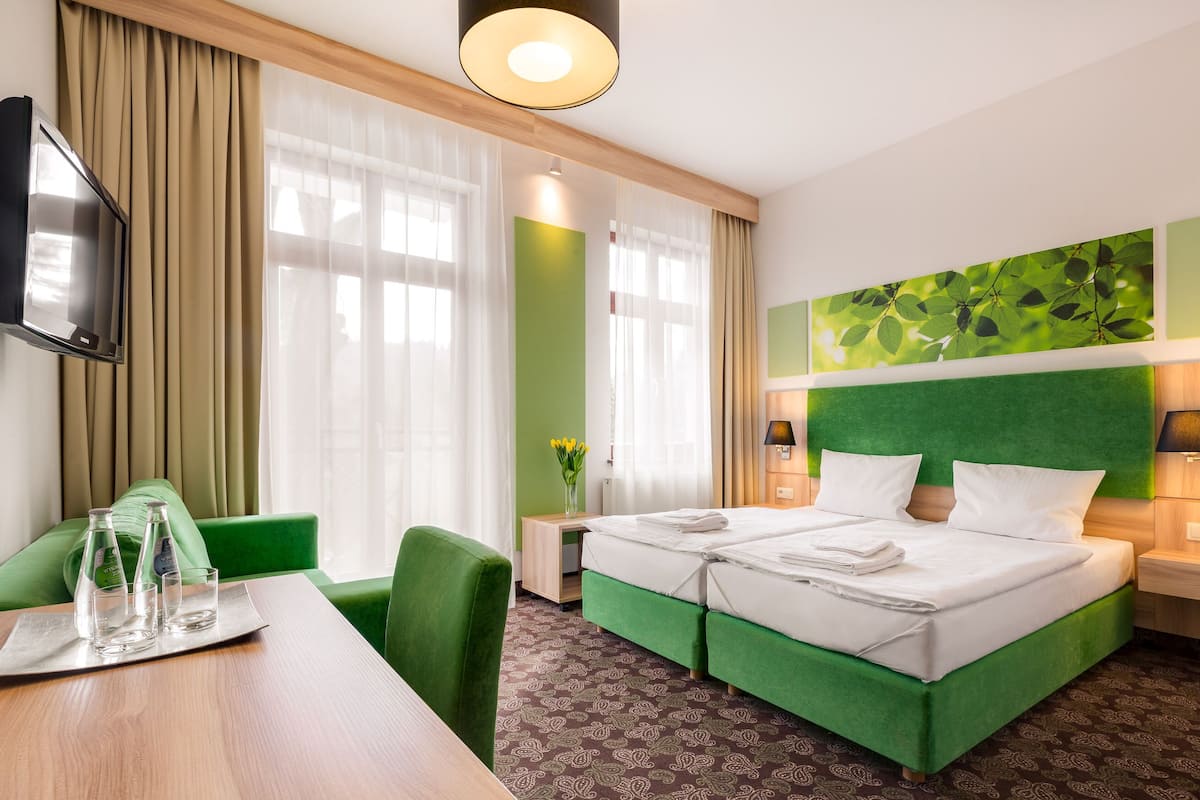Camera d'albergo con un letto imbottito verde, una scrivania in legno con un bicchiere d'acqua, una televisione a schermo piatto e un'ampia finestra con tende trasparenti.