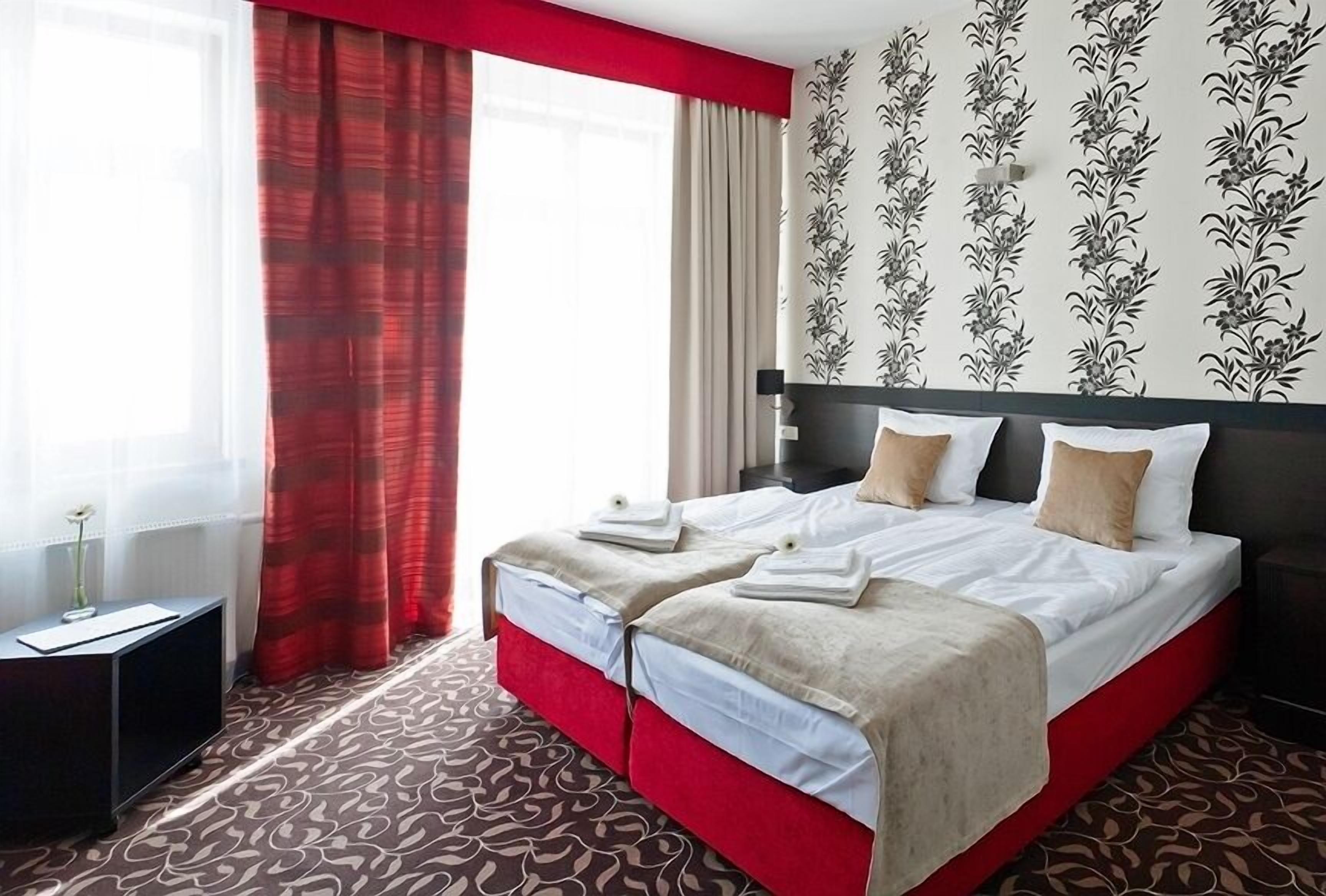 A hotel room with a large bed, red curtains, floral wallpaper, and a patterned carpet.