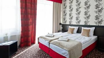 A hotel room with a large bed, red curtains, floral wallpaper, and a patterned carpet.