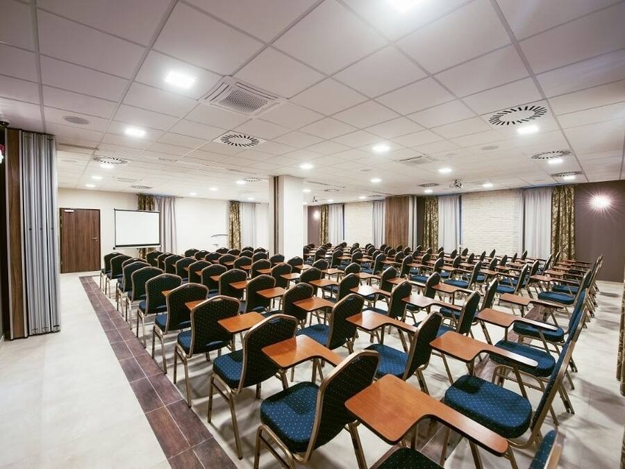 Uma sala de conferências com fileiras de mesas e cadeiras, um quadro branco e uma tela de projetor.
