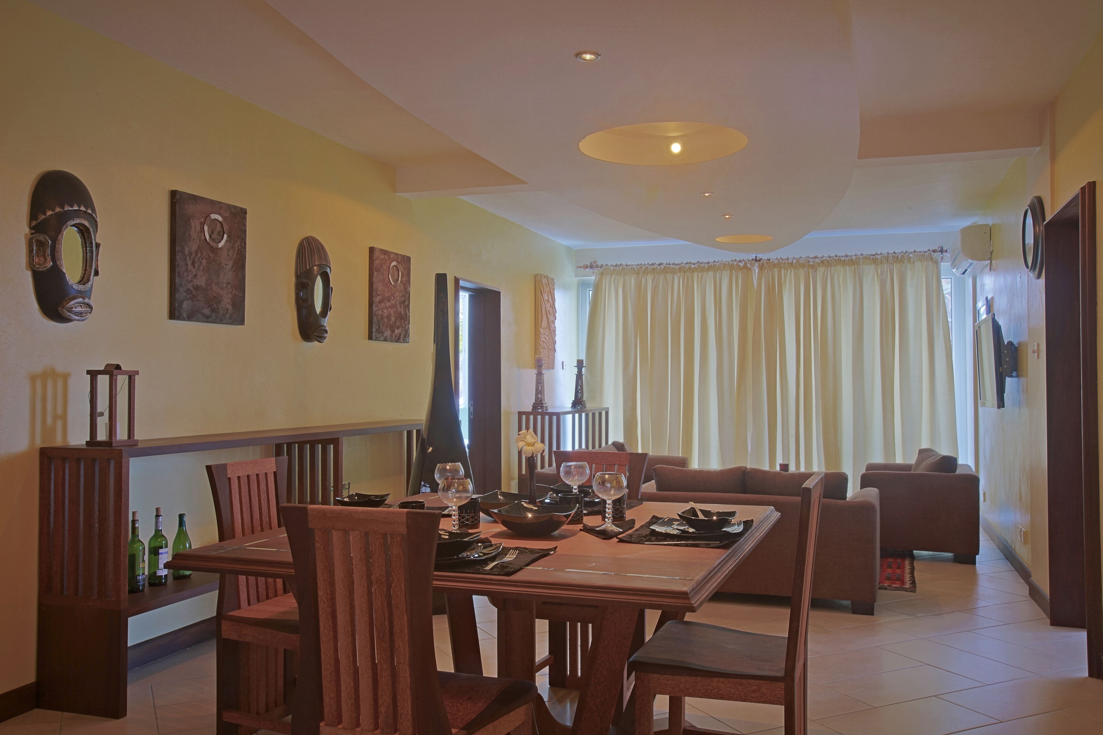 Un coin repas avec une table en bois dressée pour quatre personnes, entourée de chaises. On peut voir des masques décoratifs accrochés au mur, un canapé au fond et une fenêtre avec des rideaux.