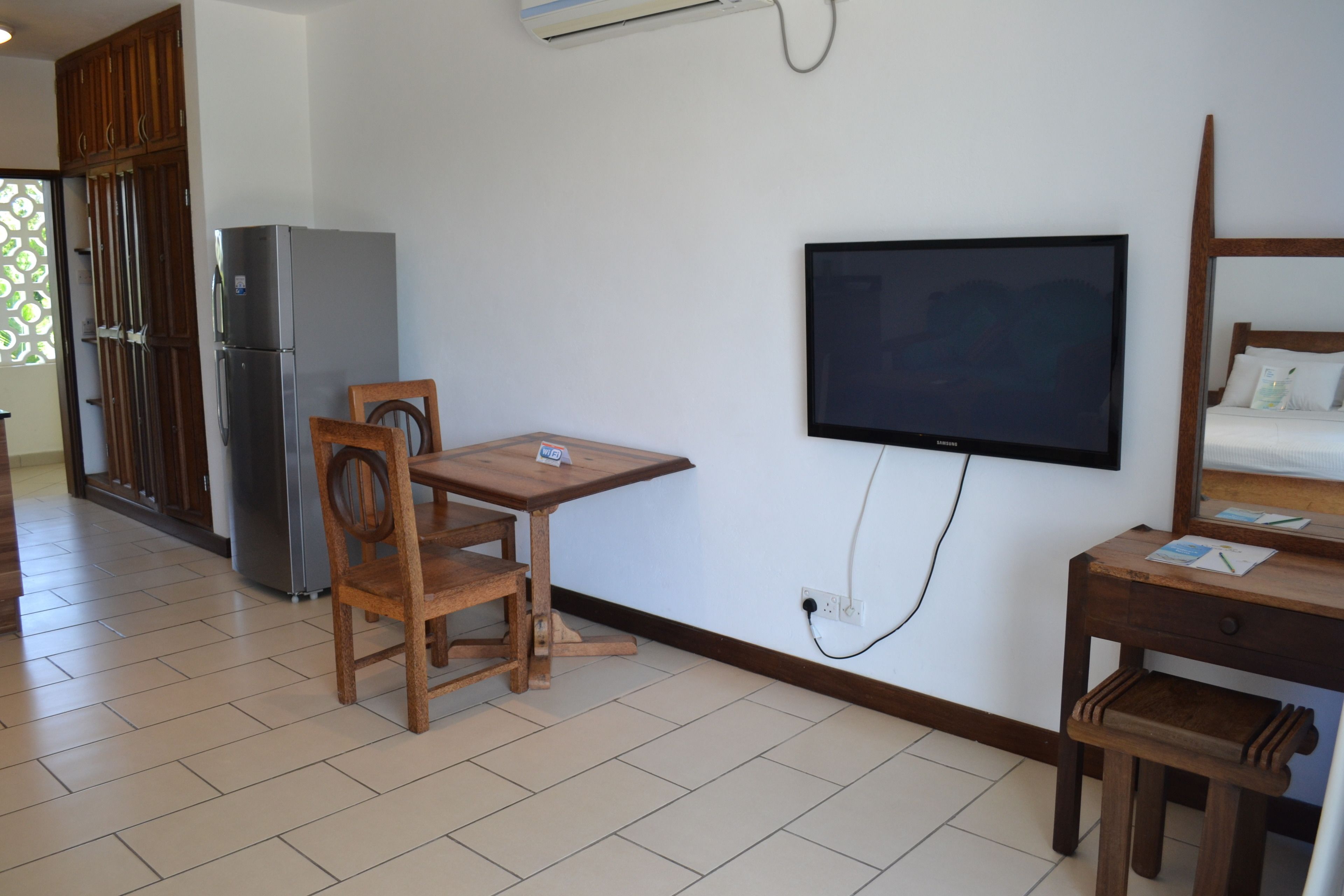 A compact room with a wooden dining table, chairs, a flat-screen TV mounted on the wall, a small wooden desk, and a refrigerator.
