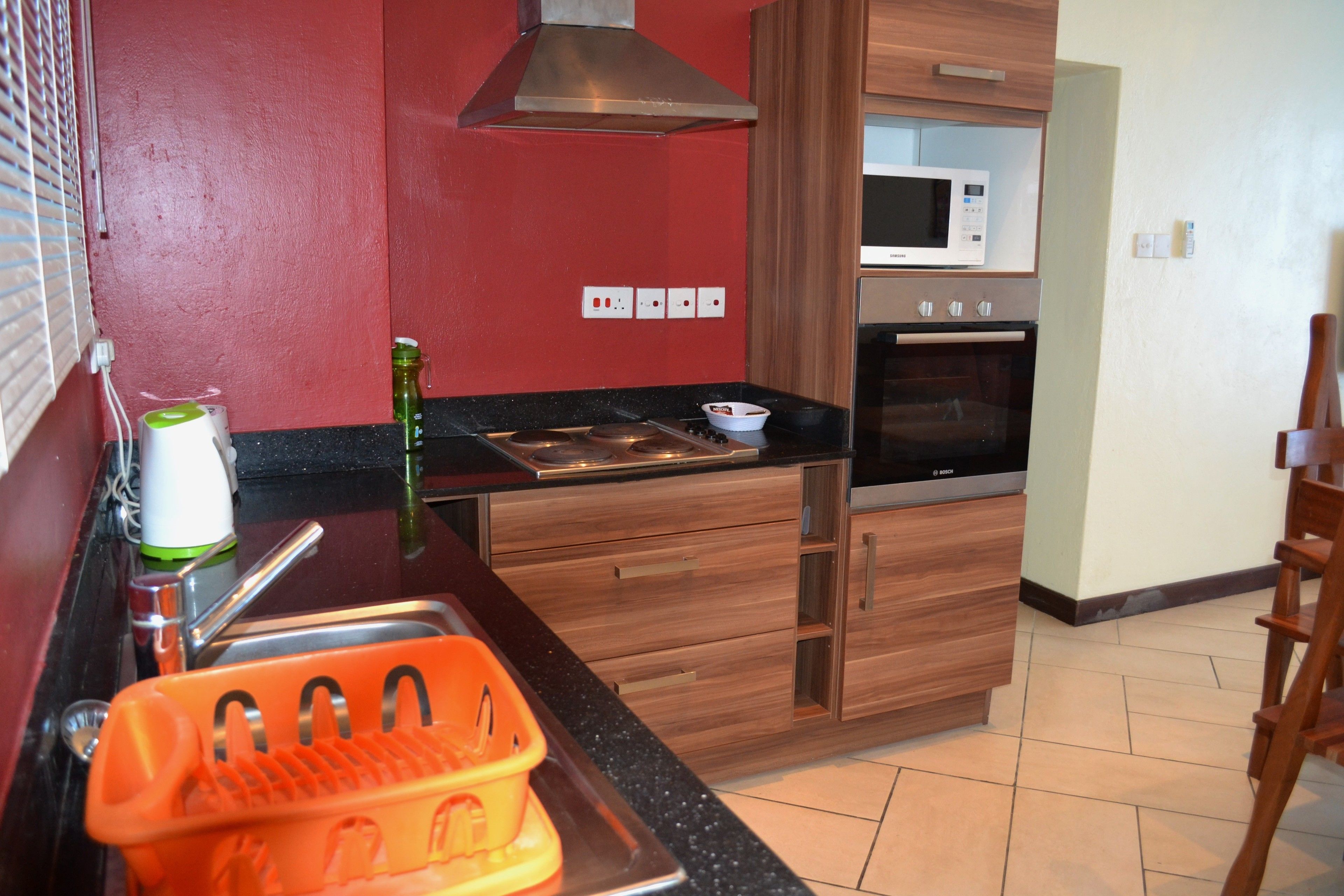 Una cocina con microondas, horno y armarios de madera. Hay un lavamanos con un escurreplatos y una botella verde en la encimera.
