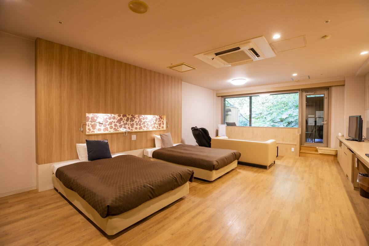 A modern hotel room with two beds, a small seating area, a TV, and a large window offering a view of greenery.
