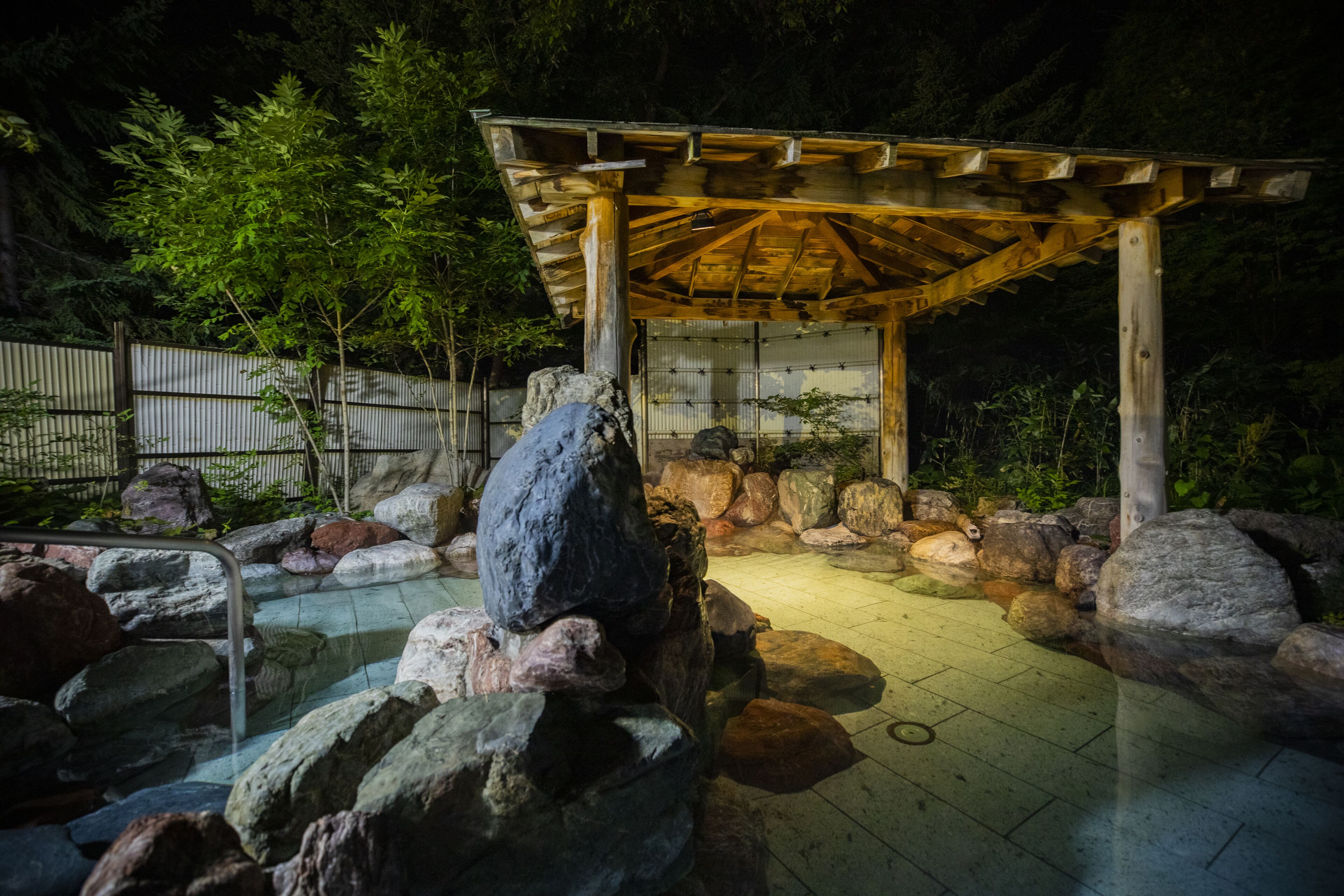 Een houten pergola met betegelde vloer, omringd door rotsen en een klein vijvertje.