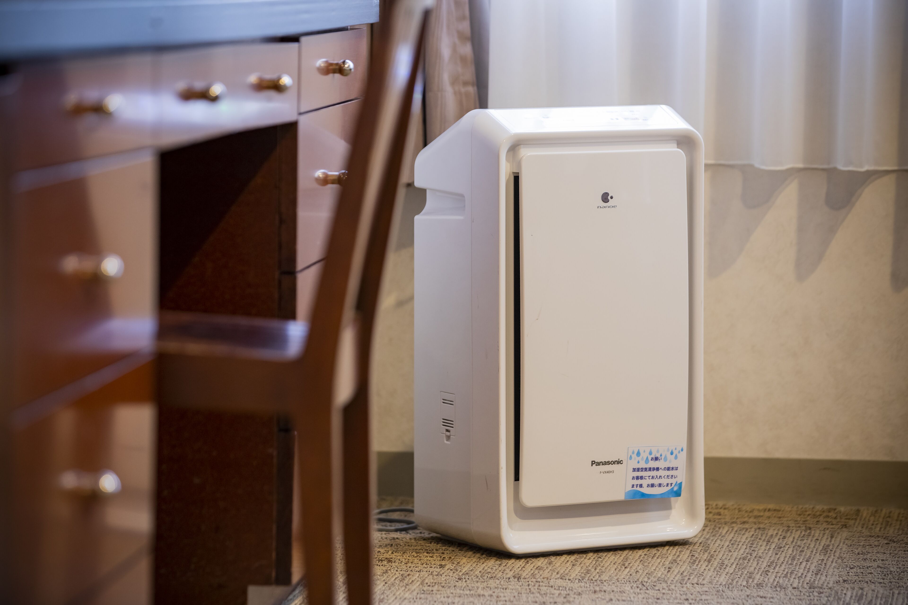 A white Panasonic refrigerator in a hotel room with a wooden dresser and a curtain in the background.